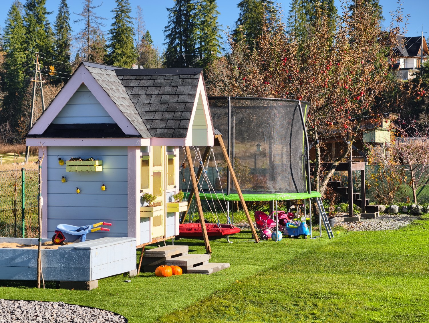 Plac zabaw w ogrodzie. Huśtawki, piaskownica z izolowanym spodem, trampolina, platforma, domek dla dzieci.