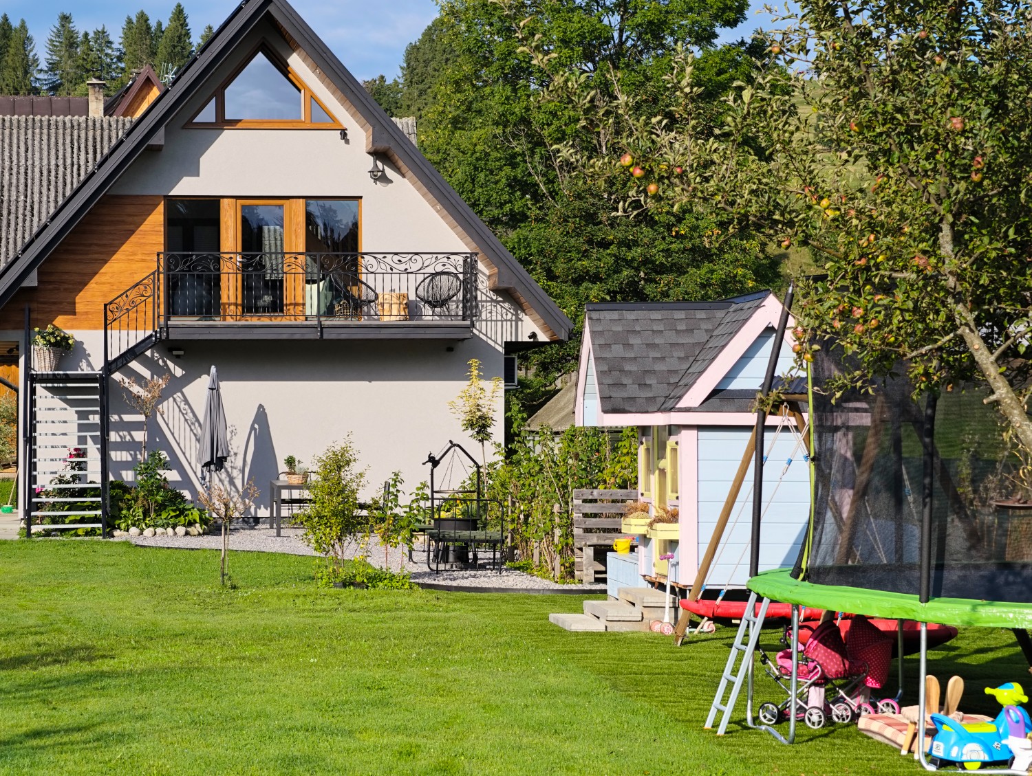 Widok na domek i fragment ogrodu. z placem zabaw. Pod domkiem duży stół, parasol, miejsce na grilla.